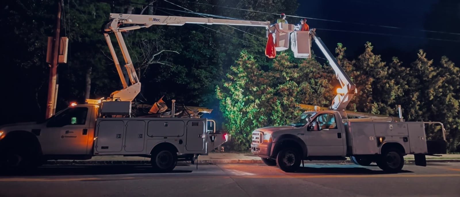 Nighttime utility work with two parked bucket trucks, elevated technicians and softly pulsing amber warning lights.